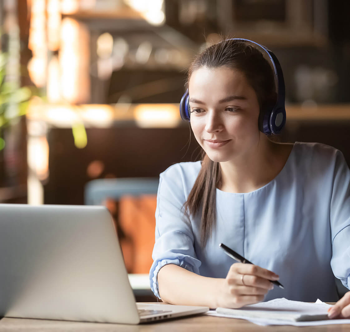 Focused woman wearing headphones using laptop, writing notes.