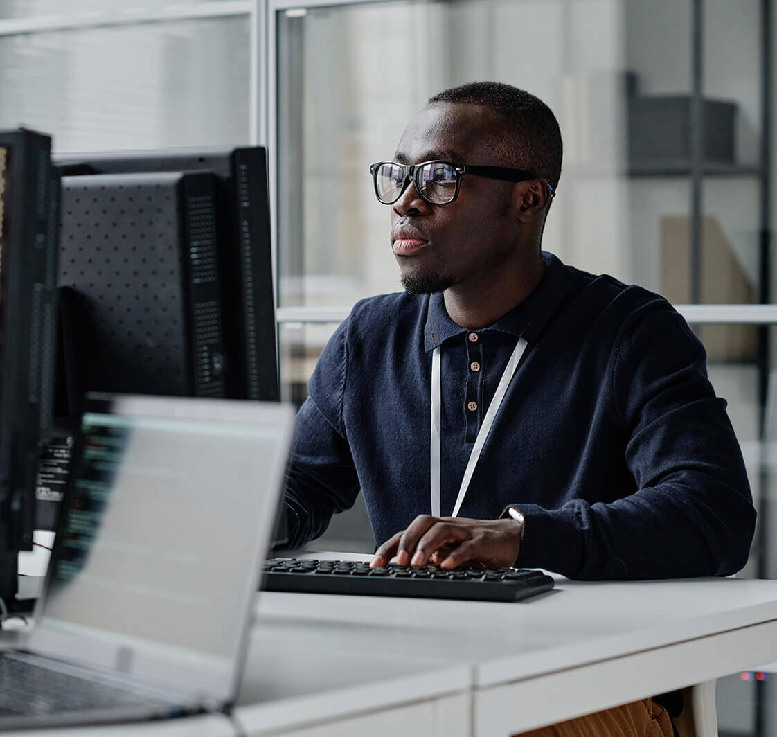 Young developer in eyeglasses concentrating on his online work on computer sitting at workplace.