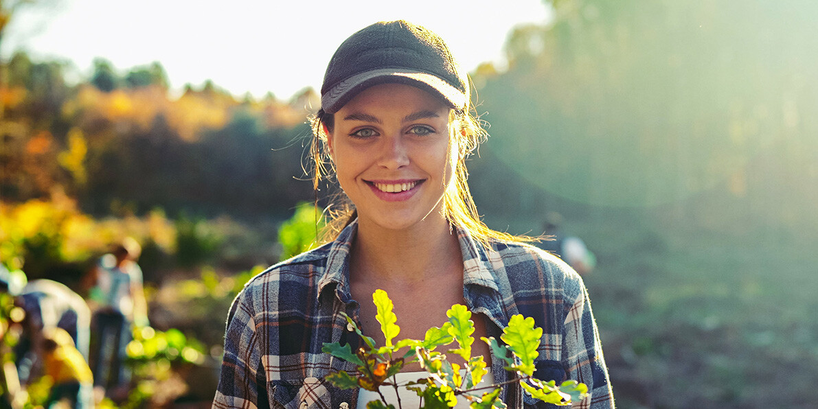 Portrait shot of young woman standing outside with tree seedling in pot and smiling cheerfully.